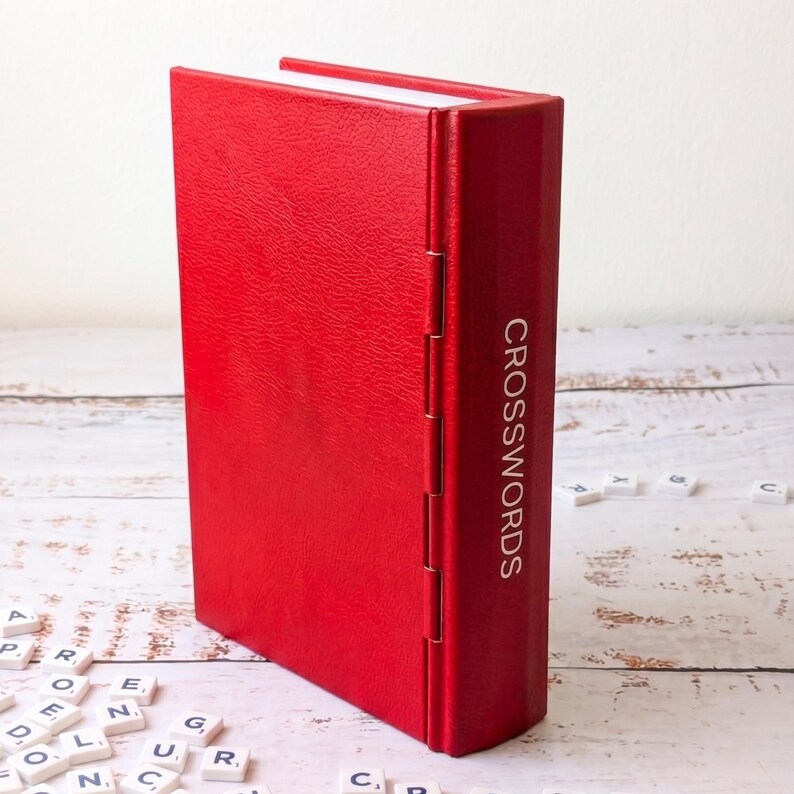 Puede incluir: Un libro rojo de tapa dura con la palabra "CROSSWORDS" impresa en el lomo. El libro est&aacute; de pie. Las fichas de Scrabble est&aacute;n esparcidas alrededor del libro, lo que sugiere un tema de crucigramas.