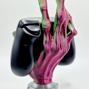 May include: A 3D printed hand-shaped game controller holder. The hand is pink and green with a silver base. The controller is black.