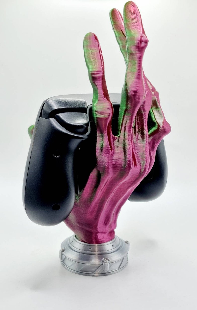 May include: A 3D printed hand-shaped game controller holder. The hand is pink and green with a silver base. The controller is black.