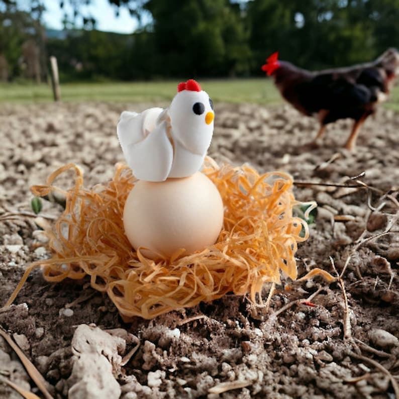May include: A white ceramic chicken figurine sits on a large brown egg in a nest of straw. The chicken has a red comb and a yellow beak. A brown hen is visible in the background.