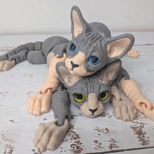 May include: Two gray and tan Sphynx cat sculptures are intertwined. The cats have blue and yellow eyes, with articulated limbs and a detailed, textured appearance. The sculptures are posed on a light-colored wooden surface.
