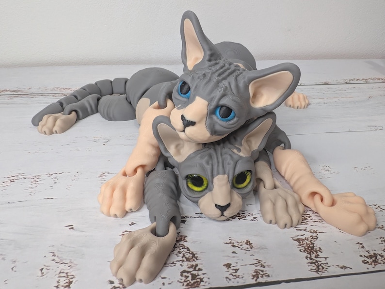 May include: Two gray and tan Sphynx cat sculptures are intertwined. The cats have blue and yellow eyes, with articulated limbs and a detailed, textured appearance. The sculptures are posed on a light-colored wooden surface.