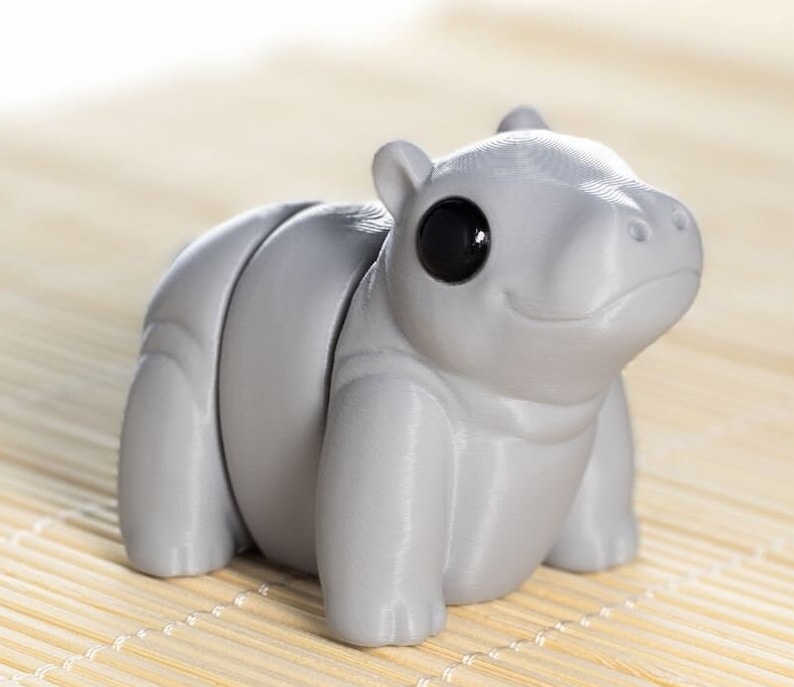 May include: A gray, 3D printed, articulated hippopotamus toy with a black eye. The toy is sitting on a bamboo mat.