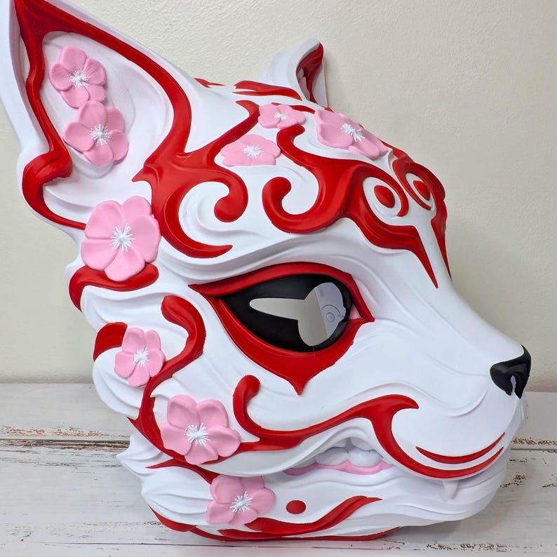 May include: A white fox mask with red accents and pink flower embellishments. The mask features a black nose and eye details, with a stylized design. The overall aesthetic is decorative and artistic.