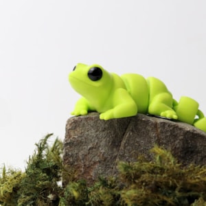 May include: A bright green frog figurine with black eyes, sitting on a gray rock covered in green moss.