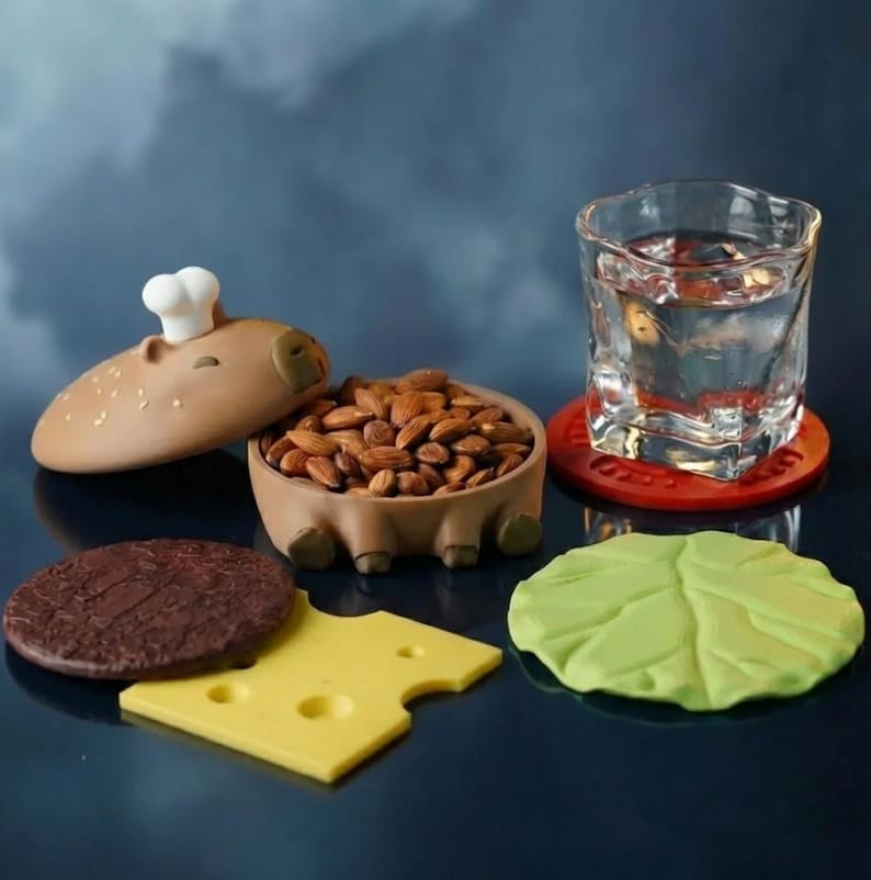 Op de afbeelding: Een grillige capibara-vormige container gevuld met amandelen, naast een glas water op een rode onderzetter. Inclusief een bruin hamburgerbroodje deksel met een koksmuts, een chocoladekleurige burger patty, een gele kaasschijf en een groen sla blad.