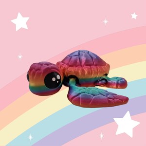 Puede incluir: Una peque&ntilde;a figura de tortuga de color arco&iacute;ris con un acabado brillante e iridiscente. La tortuga est&aacute; tumbada boca abajo con la cabeza girada hacia un lado. La figura est&aacute; sobre un fondo de arco&iacute;ris.