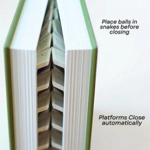 May include: A green book with white pages and a unique interior design. The text on the image reads "Place balls in snakes before closing" and "Platforms Close automatically". The book's interior features a series of platforms.