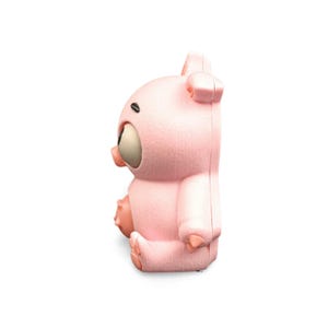 May include: A pink, cartoon-style pig figurine with large, expressive eyes and small ears. The pig is sitting, with its arms and legs visible. The figurine has a smooth, matte finish and is set against a white background.