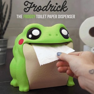 May include: A green frog-shaped toilet paper dispenser with large black and white eyes and a red cheek. The dispenser is holding a piece of white toilet paper. The text "Frodrick The Froggy Toilet Paper Dispenser" is above the frog.