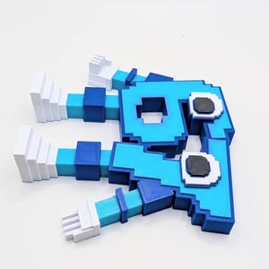 May include: A 3D-printed, pixelated figure in shades of blue, turquoise, and white. The figure has a blocky design with two arms and a head featuring two black and white eyes. The overall shape resembles a stylized creature or character.