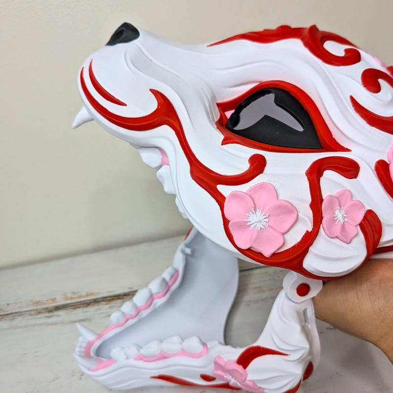 May include: A white and red decorative mask in the shape of a wolf's head. The mask features red accents, black eyes, and pink flower embellishments. The mouth is open, revealing white teeth.