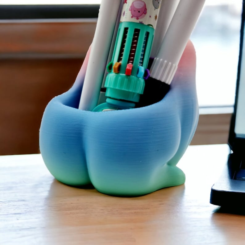 Pu&ograve; includere: Un portapenne stampato in 3D di forma unica, con una sfumatura di colori dal blu al verde. Contiene varie penne e pennarelli, tra cui una penna multicolore. Il portapenne &egrave; appoggiato su una superficie di legno.