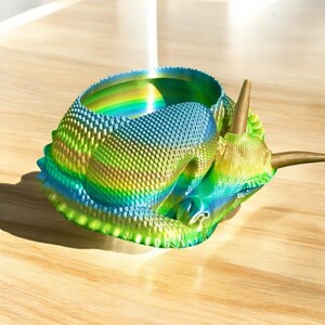 May include: A iridescent green, blue, and gold dinosaur planter with a textured surface. The planter is shaped like a triceratops and has a horn on the top.