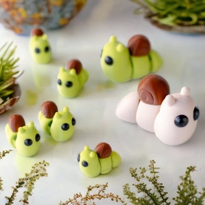 May include: A collection of handmade polymer clay snail figurines. The snails are various colors, including green, brown, and white. Some snails have brown shells, while others have green shells. The snails are all different sizes and have cute, cartoon-like faces.