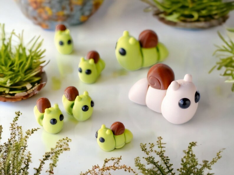May include: A collection of handmade polymer clay snail figurines. The snails are various colors, including green, brown, and white. Some snails have brown shells, while others have green shells. The snails are all different sizes and have cute, cartoon-like faces.