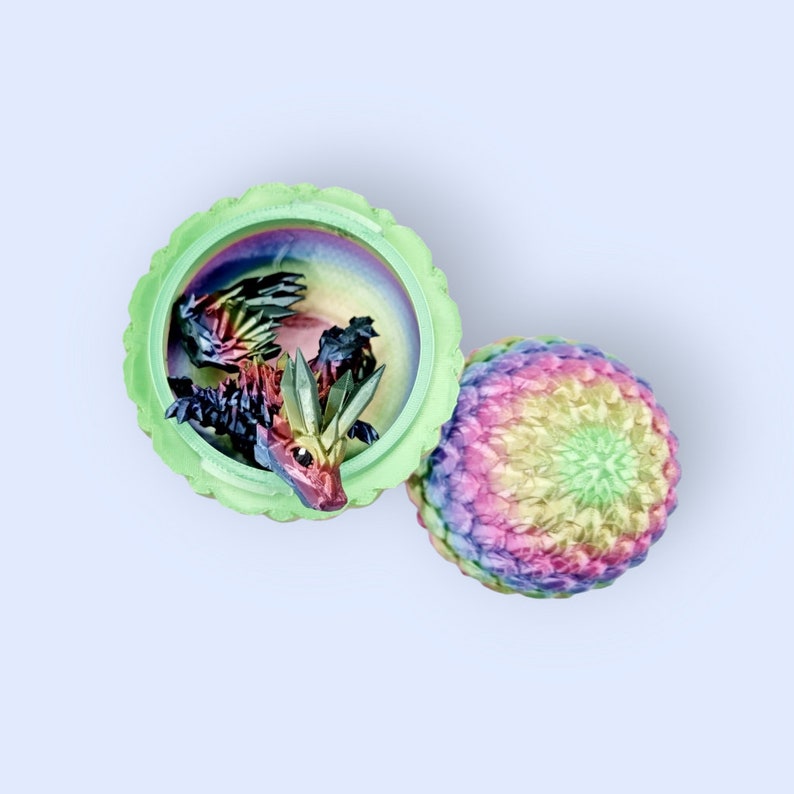 May include: A green and iridescent dragon figurine inside a green and iridescent egg-shaped container. The egg is open and the dragon is visible inside. The egg has a rainbow iridescent finish.