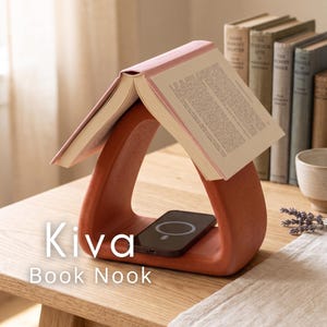 May include: A terracotta-colored book nook with a triangular design, holding an open book and a black smartphone. The text "Kiva Book Nook" is displayed on the wooden surface. The nook is designed to hold a book and charge a phone.