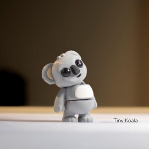 May include: A small gray koala figurine with black eyes and a white patch on its chest. The figurine is standing on a white surface. Tiny Koala is written below the figurine.