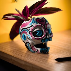 May include: A white skull-shaped planter with pink and blue floral designs, holding a plant with dark red and green leaves. The planter sits on a wooden surface against a yellow background.