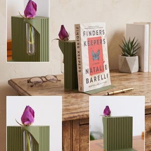 May include: A sage green metal bookend with a vertical ribbed design, holding a book titled "Finders Keepers" by Natalie Barelli. A small glass vial holds a single purple rose bud. A gold pen rests nearby.