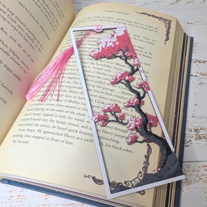 May include: A decorative bookmark featuring a cherry blossom tree design with pink blossoms and a gray trunk. The bookmark has a white border and a pink tassel. The bookmark is placed in an open book.