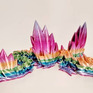 以下が含まれることがあります： 虹色の光沢のある色をした3Dプリントされたドラゴン彫刻。ドラゴンは頭、胴体、翼など、複数の部分で構成されています。ドラゴンは頭を上げた休息姿勢をとっています。