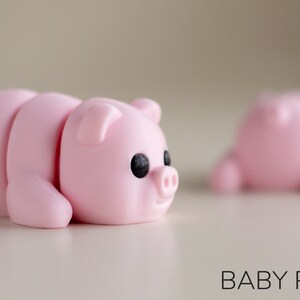 Puede incluir: Figurita de cerdo rosa claro segmentada, con ojos negros y un peque&ntilde;o hocico. La figurita est&aacute; tumbada, con un segundo cerdo borroso en el fondo. El texto "BABY PIG" est&aacute; en la parte inferior.