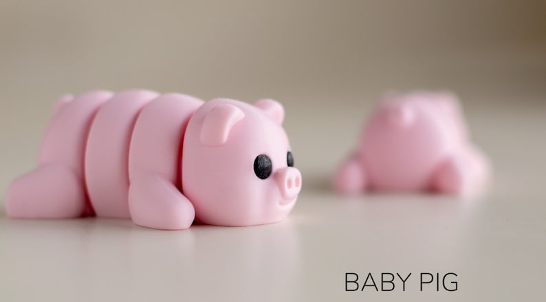 Puede incluir: Figurita de cerdo rosa claro segmentada, con ojos negros y un peque&ntilde;o hocico. La figurita est&aacute; tumbada, con un segundo cerdo borroso en el fondo. El texto "BABY PIG" est&aacute; en la parte inferior.