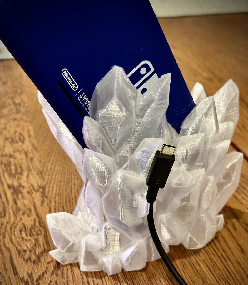 May include: A white 3D printed crystal-shaped stand for a Nintendo Switch console. The console is blue and is partially inserted into the stand. A black USB-C cable is plugged into the console and hangs down from the stand.