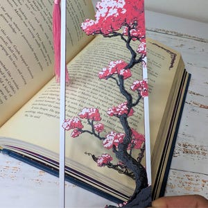 May include: A decorative bookmark featuring a cherry blossom tree design with pink flowers and a dark gray trunk. The bookmark has a pink tassel and is placed in an open book with text visible.
