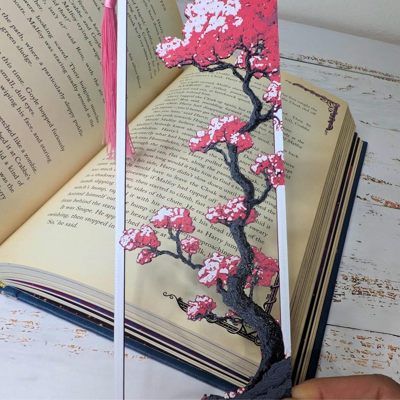 May include: A decorative bookmark featuring a cherry blossom tree design with pink flowers and a dark gray trunk. The bookmark has a pink tassel and is placed in an open book with text visible.
