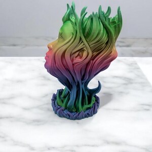 K&ouml;nnte beinhalten: Eine regenbogenfarbene 3D-gedruckte Skulptur eines Gesichts mit nach unten flie&szlig;endem Haar, das eine Basis bildet. Die Skulptur ist aus Kunststoff und hat eine glatte Oberfl&auml;che.