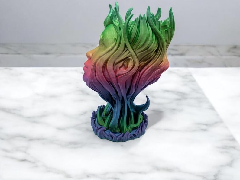 K&ouml;nnte beinhalten: Eine regenbogenfarbene 3D-gedruckte Skulptur eines Gesichts mit nach unten flie&szlig;endem Haar, das eine Basis bildet. Die Skulptur ist aus Kunststoff und hat eine glatte Oberfl&auml;che.