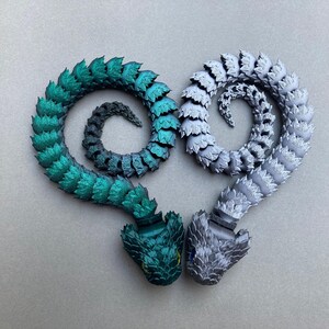 May include: Two articulated snake-like figures, one green and one silver, are curled into a heart shape. The figures are made of plastic and have a scaly texture.