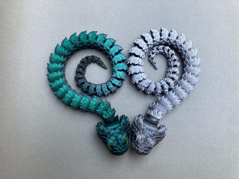 May include: Two articulated snake-like figures, one green and one silver, are curled into a heart shape. The figures are made of plastic and have a scaly texture.