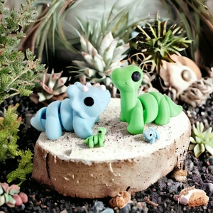 Puede incluir: Dos peque&ntilde;as figuritas de dinosaurios pintadas a mano. Una es un triceratops azul y la otra un estegosaurio verde. Ambas est&aacute;n sentadas sobre una superficie de madera.