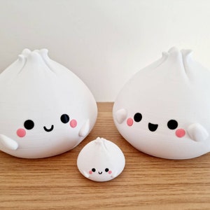 May include: Three white ceramic figurines shaped like steamed buns with smiling faces and pink cheeks. The buns are different sizes, with the smallest bun in the front and the two larger buns behind it.