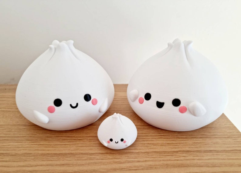 May include: Three white ceramic figurines shaped like steamed buns with smiling faces and pink cheeks. The buns are different sizes, with the smallest bun in the front and the two larger buns behind it.