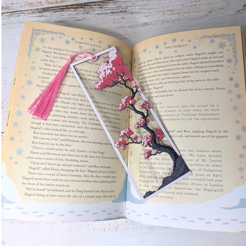 May include: A decorative bookmark featuring a cherry blossom design with pink flowers and a black branch, set against a white frame. A pink tassel hangs from the top. The bookmark is placed in an open book.