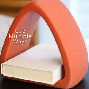 May include: An orange, arched book holder with the text "Use Multiple Ways" on the side. A closed book with cream-colored pages rests inside the holder. The holder is made of a textured material.
