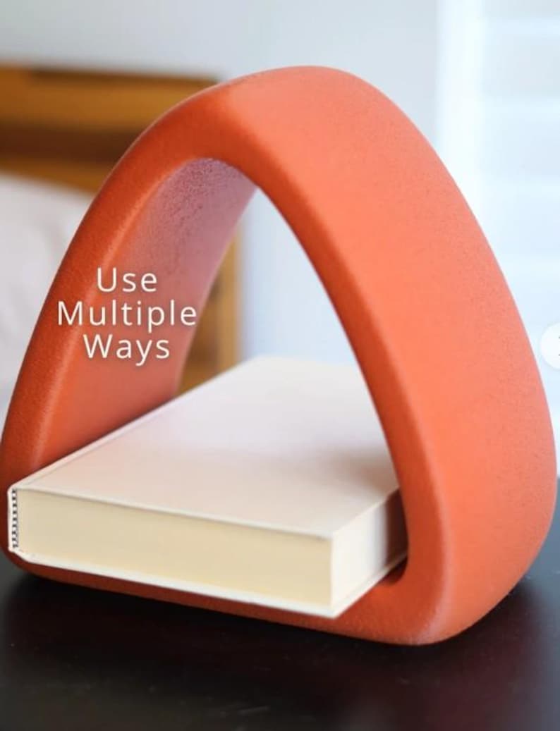 May include: An orange, arched book holder with the text "Use Multiple Ways" on the side. A closed book with cream-colored pages rests inside the holder. The holder is made of a textured material.