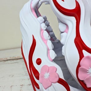 May include: Close-up of a white decorative object with red accents and a pink flower. The object has a curved shape with a series of white and gray elements, and a red circular detail. A pink flower with white details is also visible.