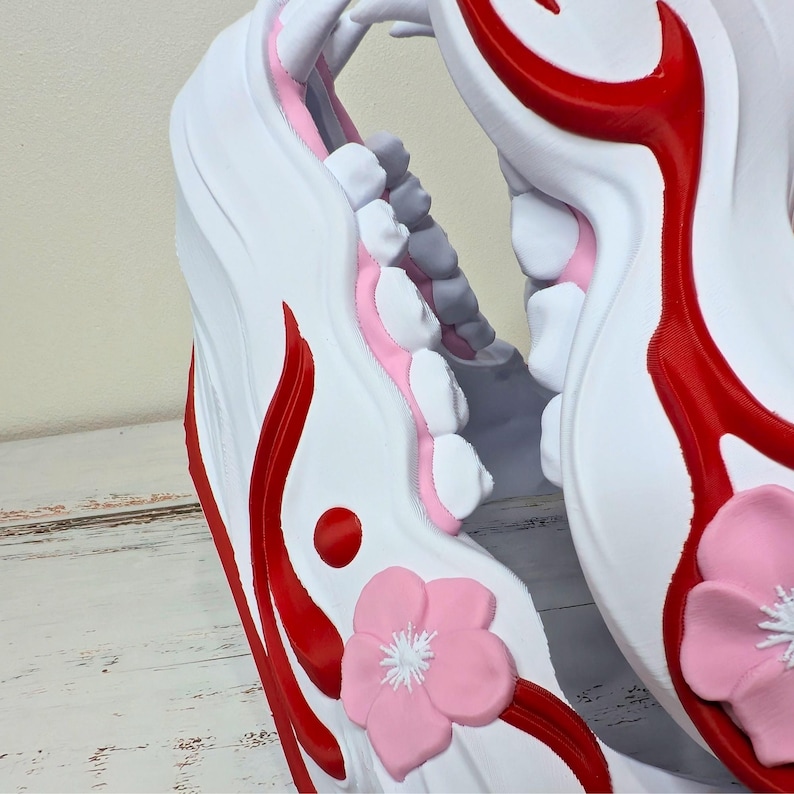 May include: Close-up of a white decorative object with red accents and a pink flower. The object has a curved shape with a series of white and gray elements, and a red circular detail. A pink flower with white details is also visible.