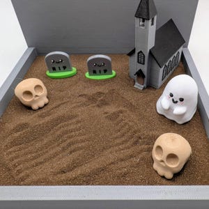 Pu&ograve; includere: Una scena di cimitero di Halloween in miniatura con una piccola chiesa, due lapidi grigie con scritto RIP, due teschi bianchi e un fantasma bianco con un viso felice.