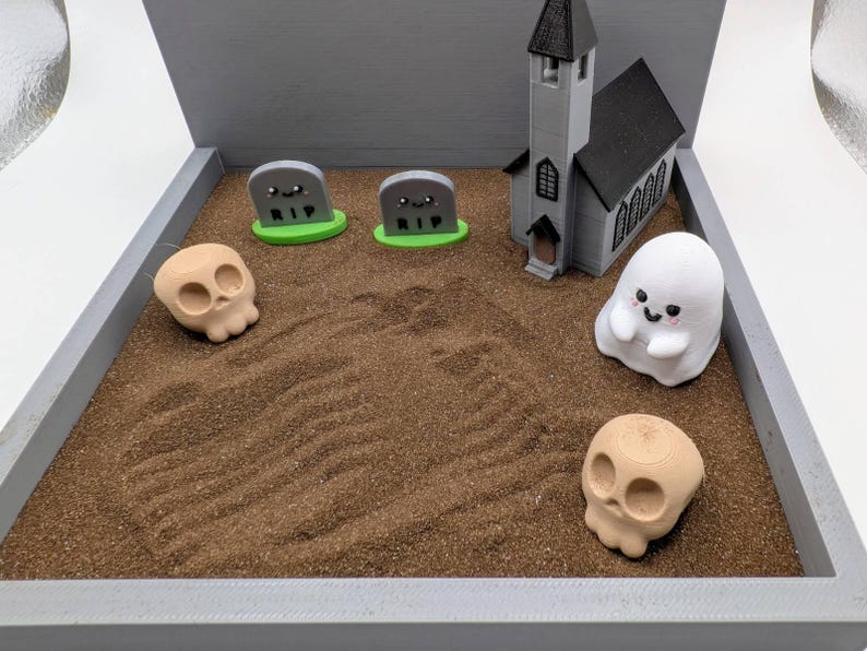 Pu&ograve; includere: Una scena di cimitero di Halloween in miniatura con una piccola chiesa, due lapidi grigie con scritto RIP, due teschi bianchi e un fantasma bianco con un viso felice.