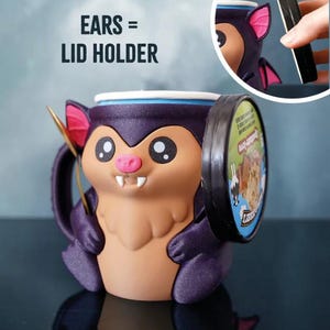May include: A novelty bat-shaped mug with a lid holder feature. The mug is primarily purple, brown, and pink, with large cartoon eyes and a smiling face. The text "EARS = LID HOLDER" is visible.