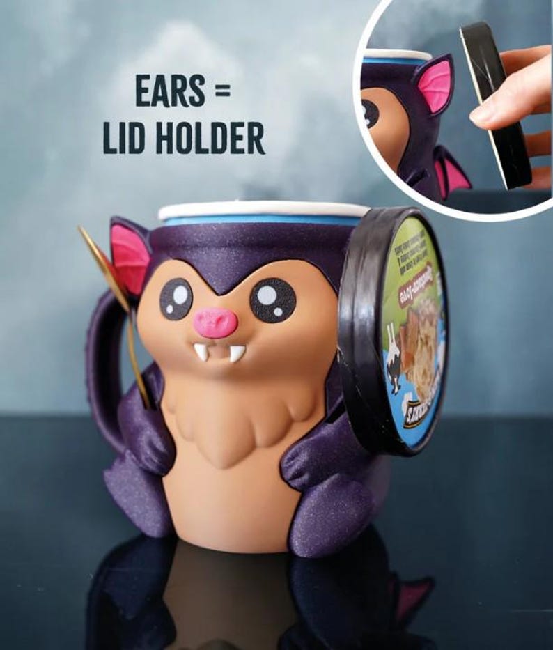 May include: A novelty bat-shaped mug with a lid holder feature. The mug is primarily purple, brown, and pink, with large cartoon eyes and a smiling face. The text "EARS = LID HOLDER" is visible.