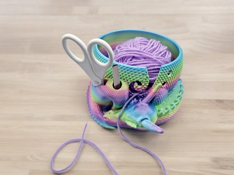 May include: A rainbow colored yarn bowl with a purple ball of yarn inside. A pair of white scissors is in the bowl. The yarn bowl is shaped like a unicorn.