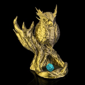 Op de afbeelding: Een goudkleurig uilbeeldje dat op een tak zit, met een turquoise, veelzijdige dobbelsteen in de basis. De uil heeft gedetailleerde veertjesstructuren en staat tegen een zwarte achtergrond. Het beeldje is waarschijnlijk een decoratief item of een dobbelsteenhouder.